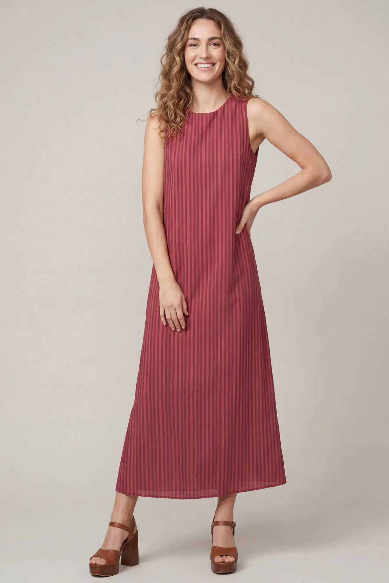 A woman modeling a sleeveless, high-neck midi dress with vertical pleats in a dusty rose color, paired with brown platform sandals, against a plain beige studio background.