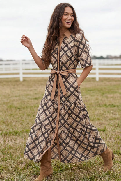 A woman wearing a beige, brown, and plaid midi dress with a tan tie belt, standing in a grassy field with a white fence in the background.
