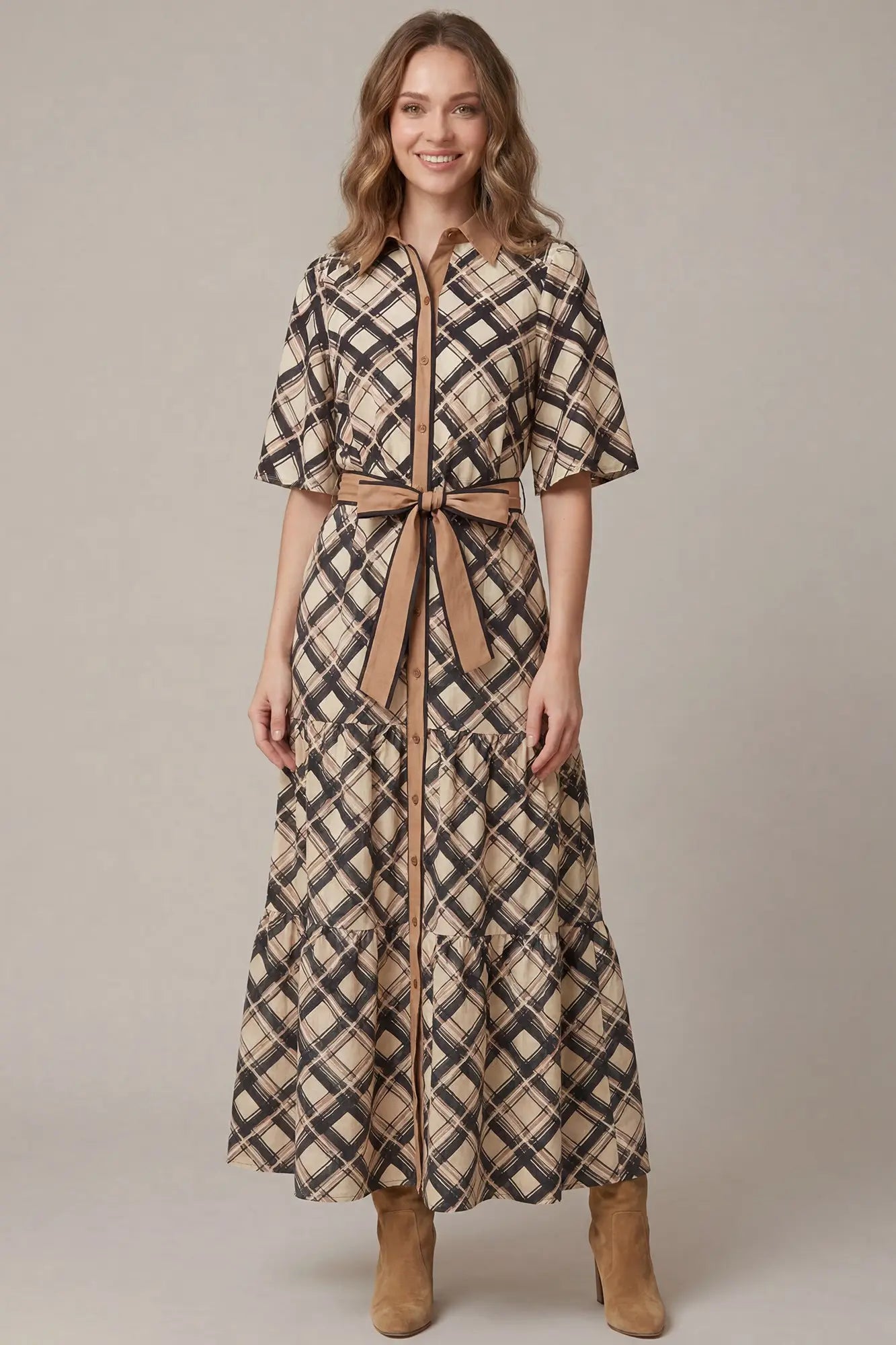 A woman modeling a long plaid shirt dress with short sleeves, a button front, and a matching waist tie, paired with tan ankle boots against a plain beige studio background.