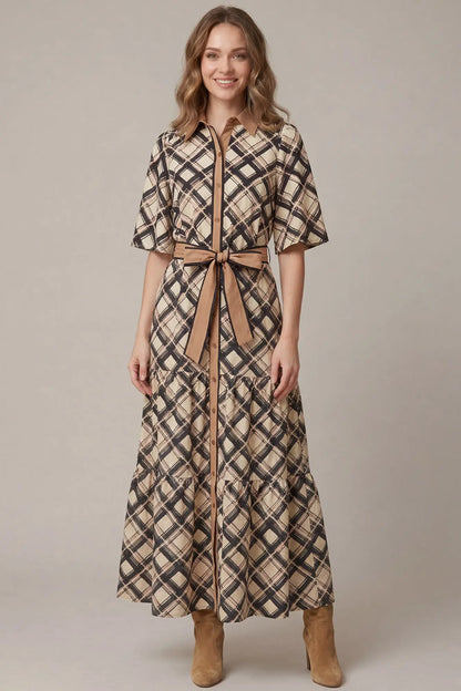 A woman modeling a long plaid shirt dress with short sleeves, a button front, and a matching waist tie, paired with tan ankle boots against a plain beige studio background.