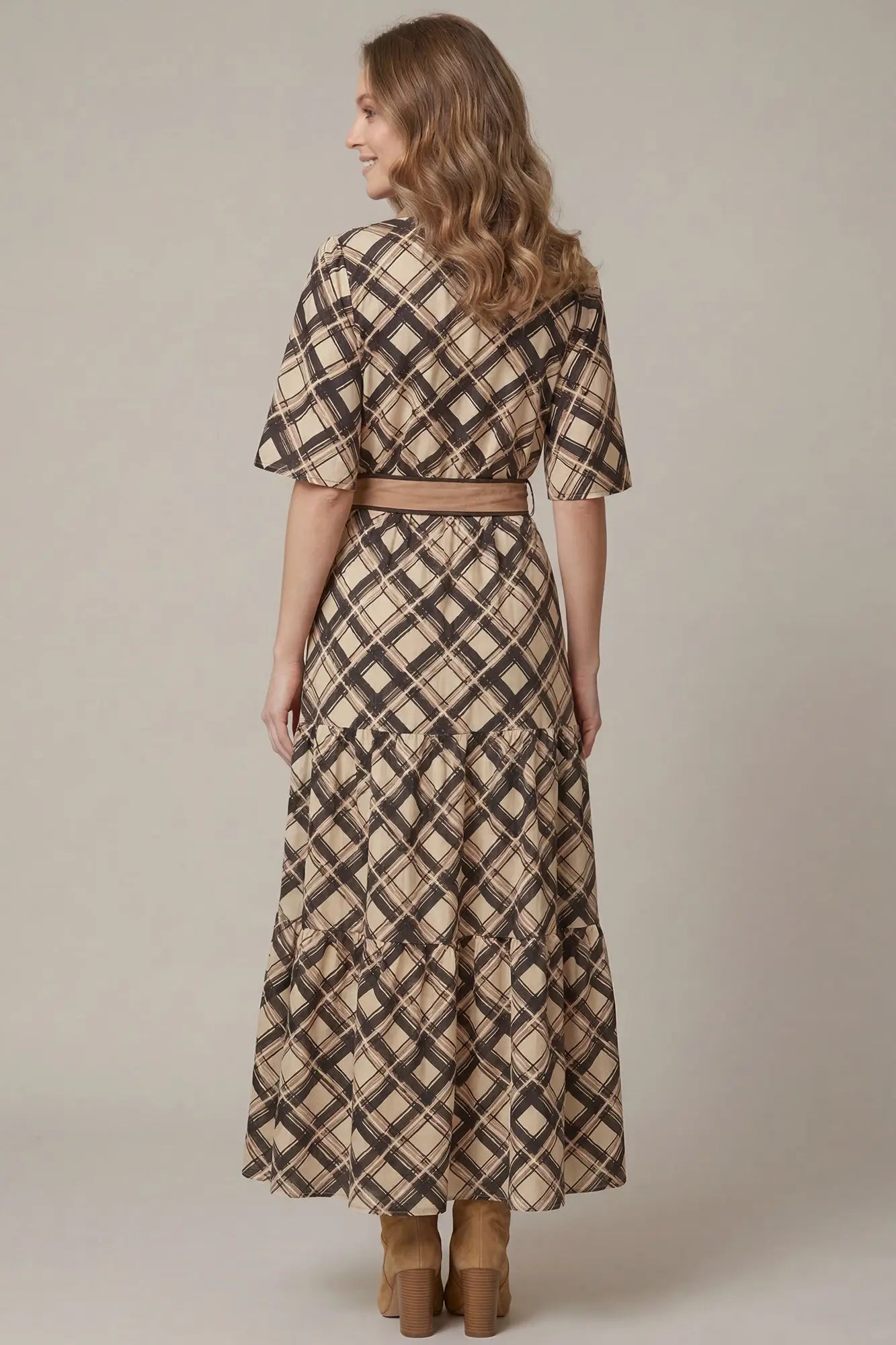 A woman shown from the back wearing a beige, brown and black plaid midi dress with short flutter sleeves and a matching belt, paired with brown boots, against a plain beige studio background.