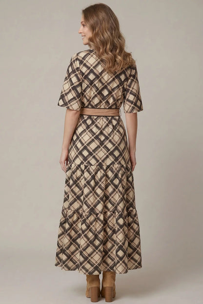 A woman shown from the back wearing a beige, brown and black plaid midi dress with short flutter sleeves and a matching belt, paired with brown boots, against a plain beige studio background.