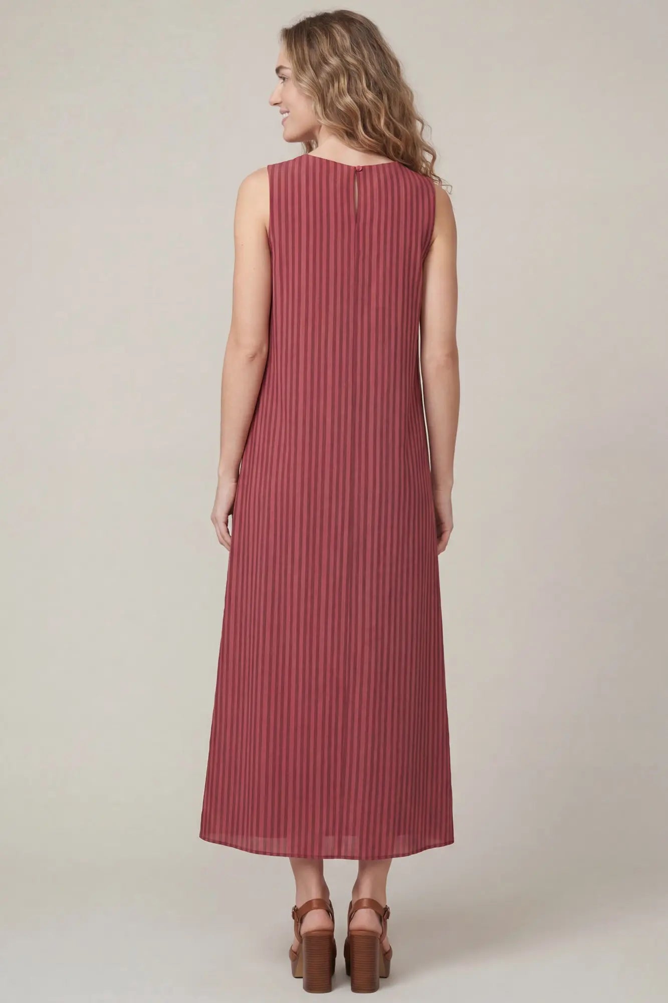 A woman wearing a sleeveless, floor-length pleated dress in a dusty rose pink color, shown from the back against a plain light beige studio background.