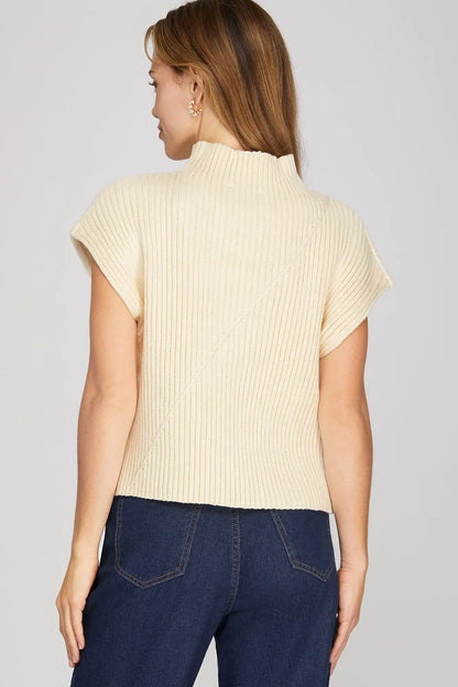 A woman shown from the back wearing a cream-colored ribbed knit short-sleeve mock neck top and dark blue jeans, set against a plain light gray studio background.
