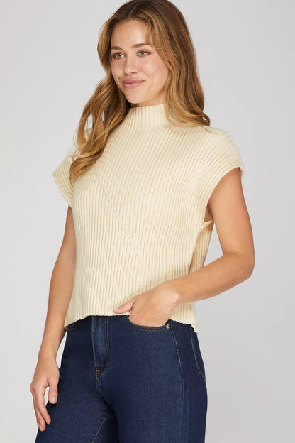 A woman wearing a cream ribbed knit mock neck top with cap sleeves and dark blue jeans, standing against a light gray studio background.