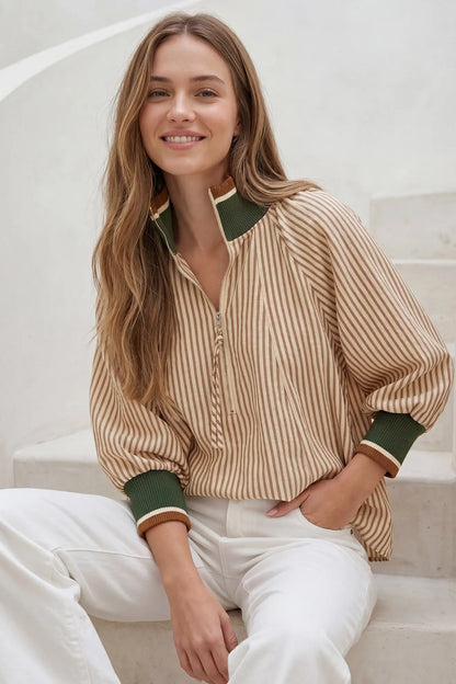 A woman sitting on white stone steps wearing a beige and white vertically striped button-up blouse with dark green ribbed collar, cuffs, and hem, paired with white pants.